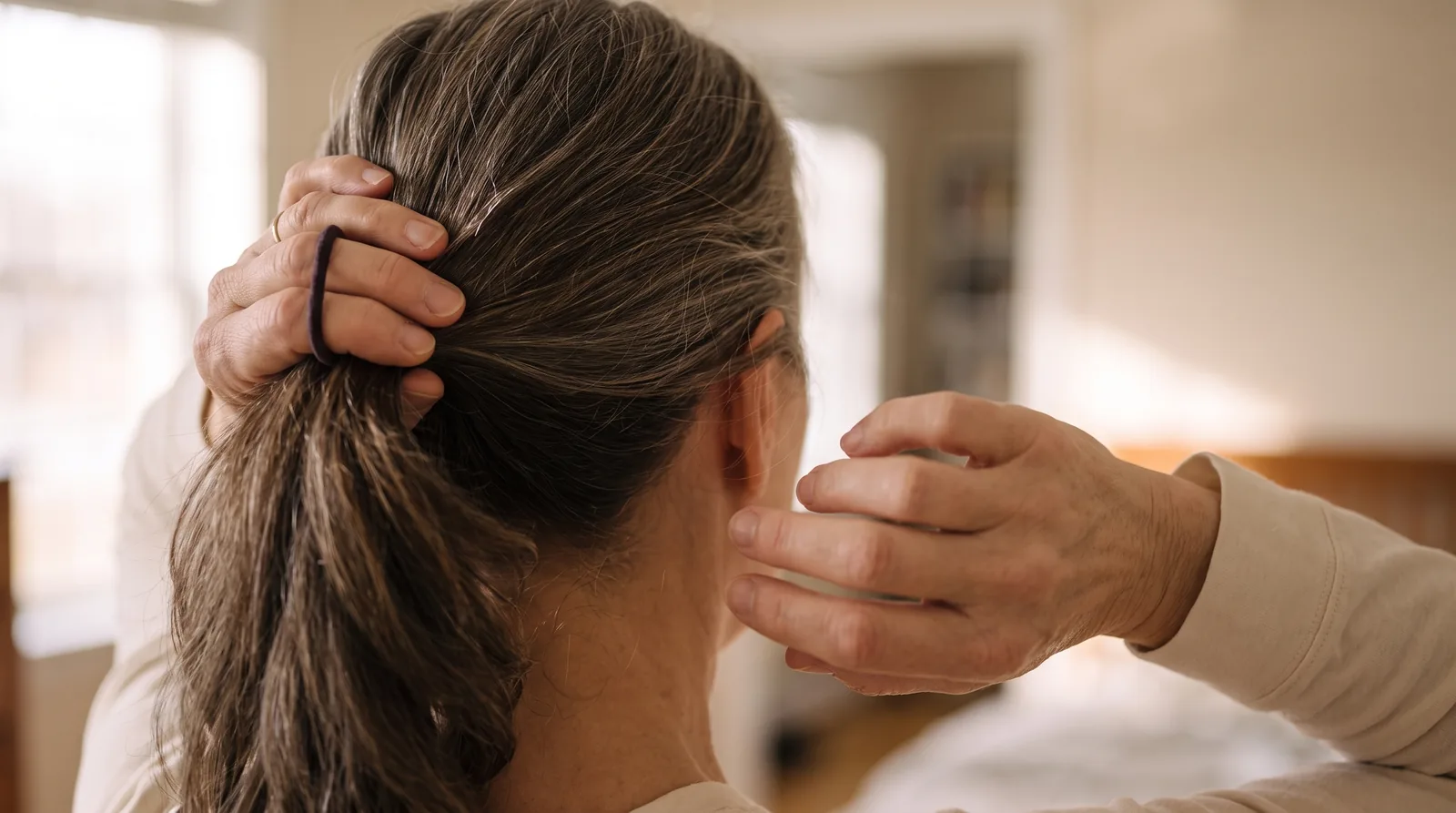 Close crop of two hands trying to gather hair at the back of the head, the right arm noticeably lower than the left